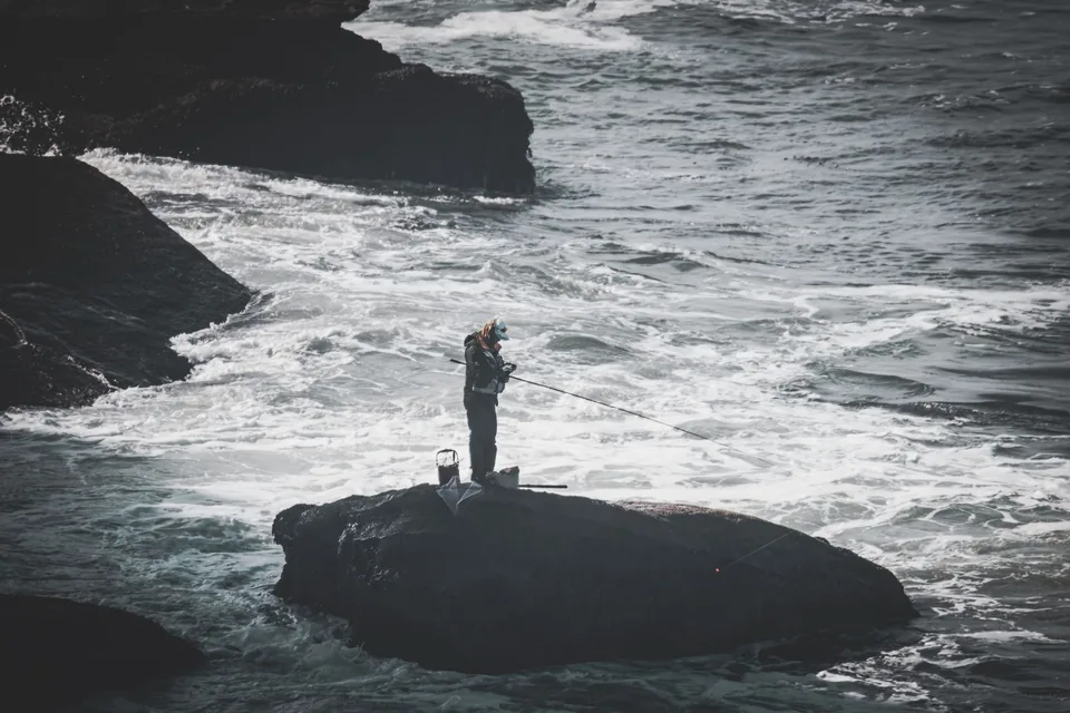 A shore angler fishes a rocky tidal edge in open coastal light
